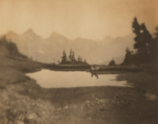 wide photograph with a wide perspective depicting a lake and mountain scene. in the distance, a nude male figure reclines next to the water