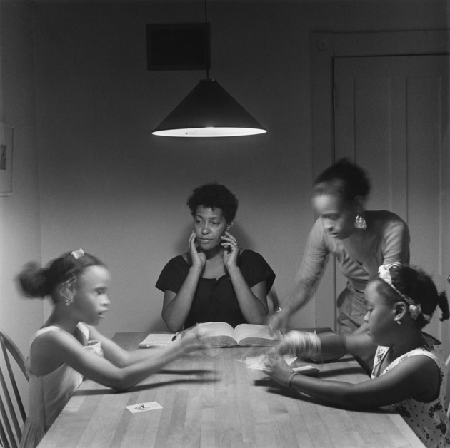 Photograph of a woman reading a book and three young girls sitting at a kitchen table playing cards