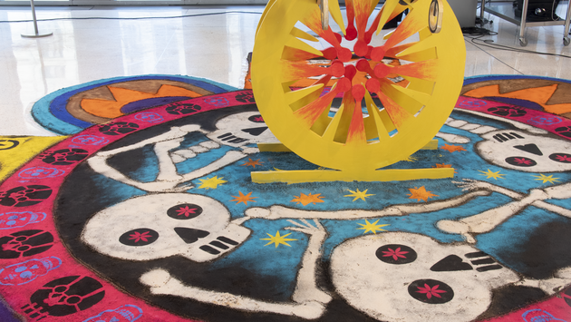 Installation photo of a tapete. Painted in the sand in the shape of a flower are skulls and stars. In the center of the tapete is a wooden structure crafted to resemble a coronavirus.