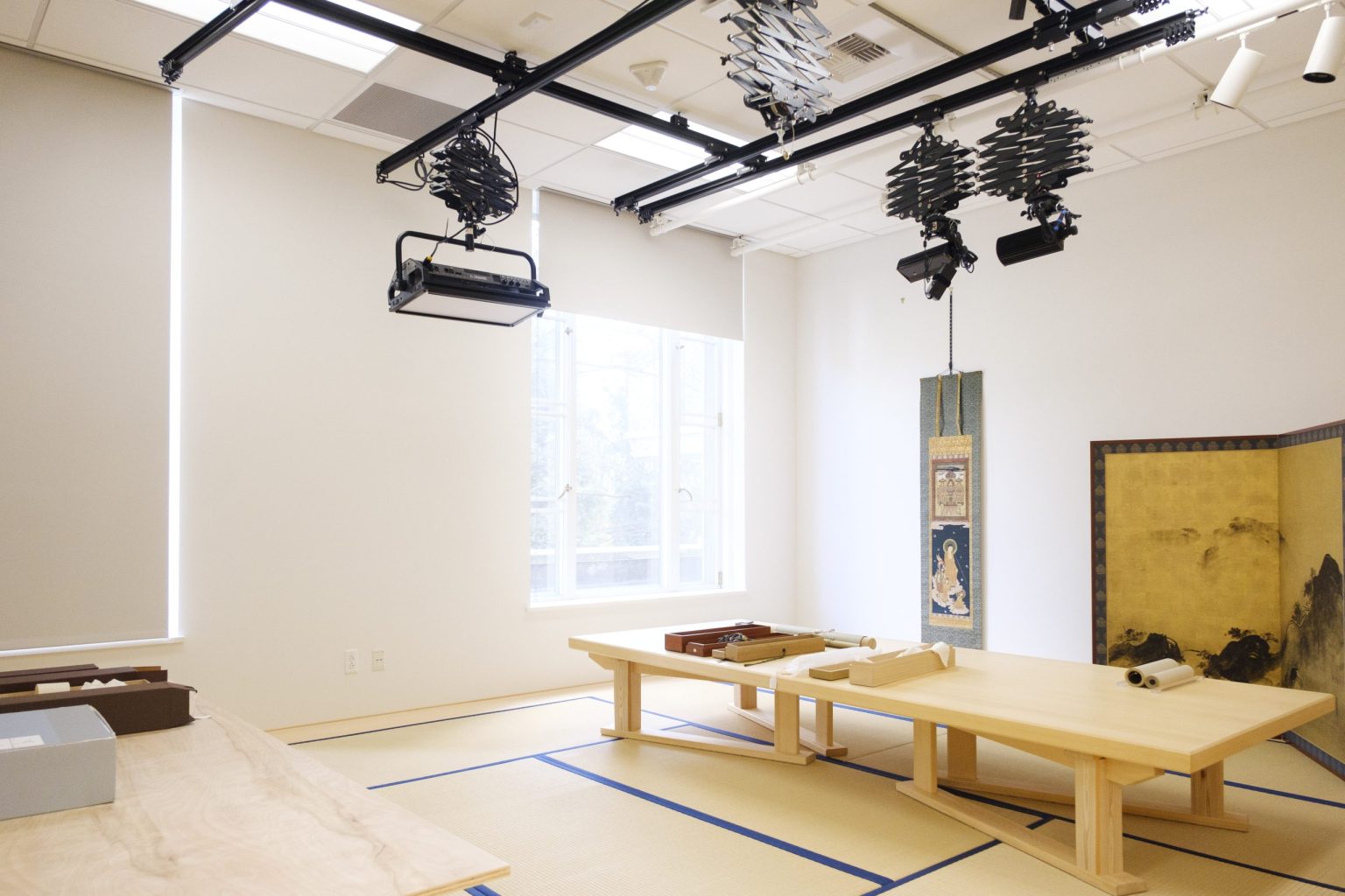 A bright, modern room with tatami mats, low wooden tables, Japanese hanging scroll art, and a decorative screen reflects Asian art and painting tradition. Ceiling-mounted lights and equipment suggest an art or restoration studio using water-based paints.