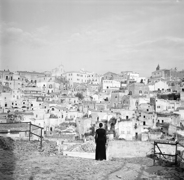 A woman in a dark gown faces away from the camera, looking out at a city and ancient buildings in white