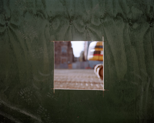 Photograph looking through a small plywood peephole at a unfocused construction site