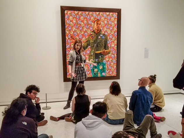 Image of a SAM staff person standing in front of a Kehinde Wiley portrait and talking to a group of students seated on the floor